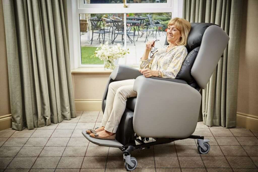 Elderly lady in Lento mobility recliner patient care chair. Indoors, the room is carpeted with beige curtains.