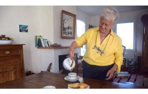 Elderly woman in her home at a table pouring a hot drink.