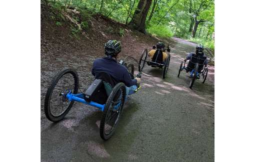 Active wheelchair users with trikes & bikes on a dirt trail in a bike park. 3 riders can be seen.
