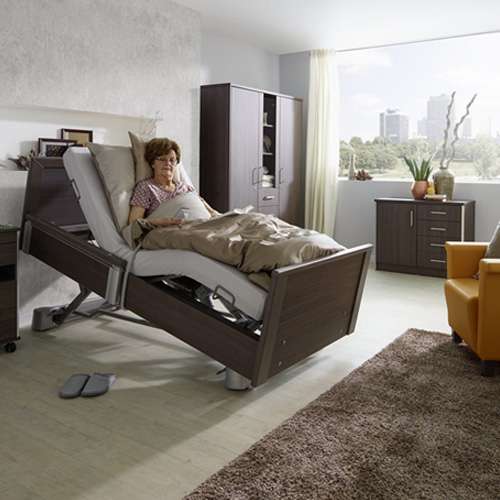 Lifestyle shot of an elderly woman in carpeted bedroom resting on a profiling bed with the headrest raised.