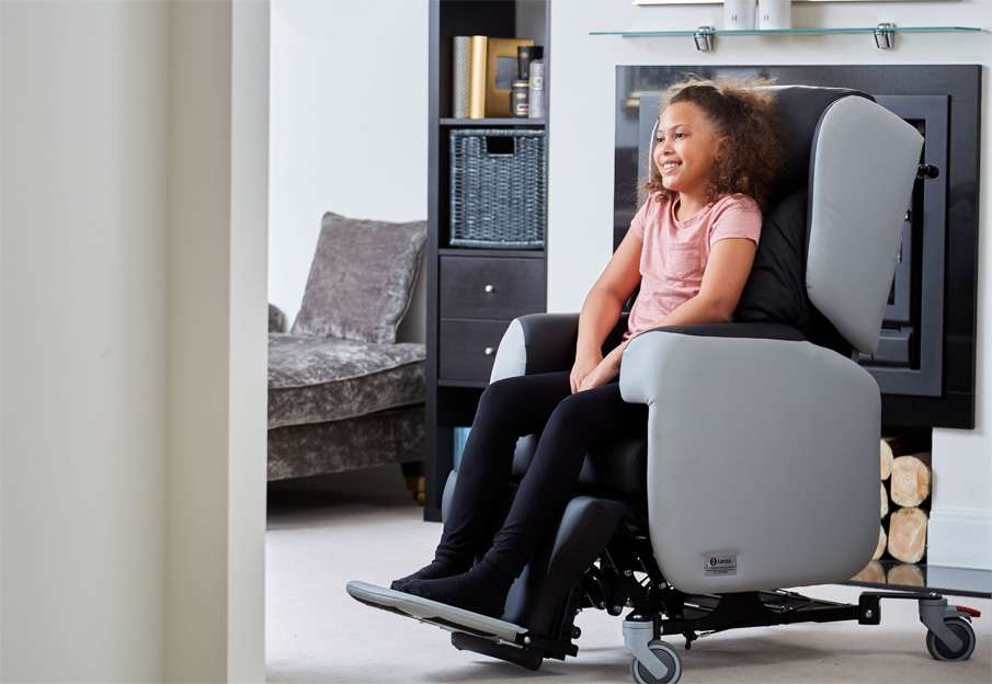 Young girl sitting in a Little Lento paediatric care chair. Indoors, in a carpeted living room.