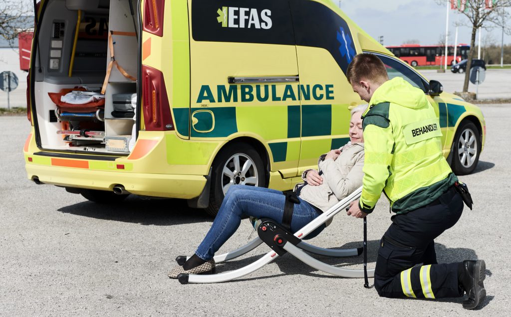 Elderly woman on a Raizer 2 lifting chair being attended to by a paramedic. They are both beside an ambulance.