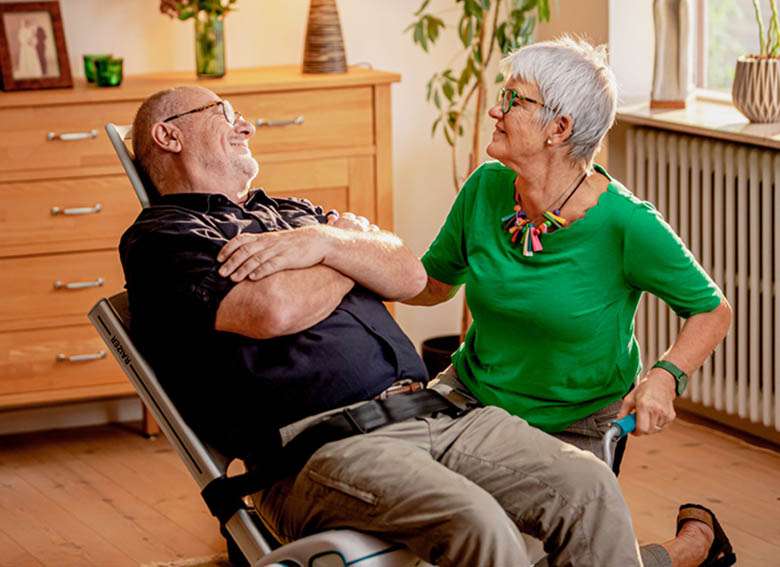 Lifestyle shot of an elderly woman helping an elderly man back to his feet using the Raizer M manual lifting chair. They are both indoors.
