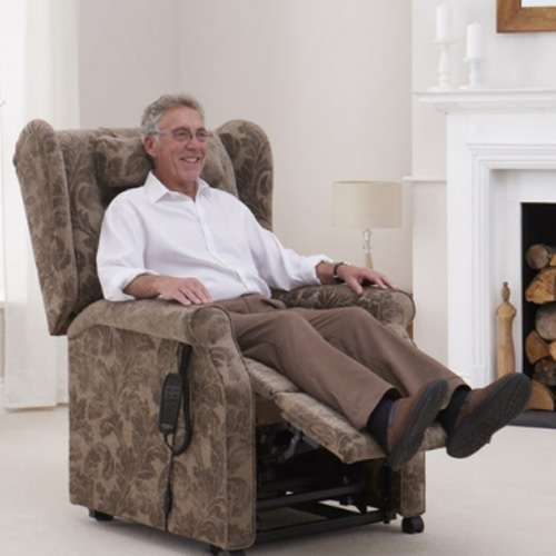 Elderly man sitting in a riser recliner armchair with the footrest up. He is indoors, there is a fireplace in the background. The carpet is cream coloured.