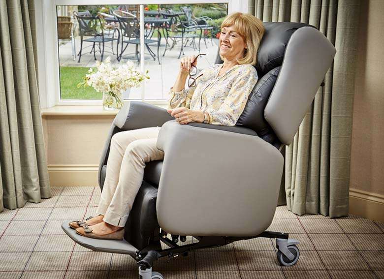 Lifestyle shot of an eldely woman sitting on a Lento adjustable care chair in her living room. Indoors and the curtains are closed.shot of a reclined lento adjustable care chair