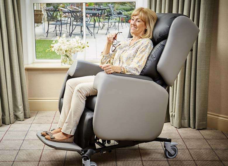 Lifestyle shot of an eldely woman sitting on a Lento adjustable care chair in her living room. Indoors and the curtains are closed.shot of a reclined lento adjustable care chair