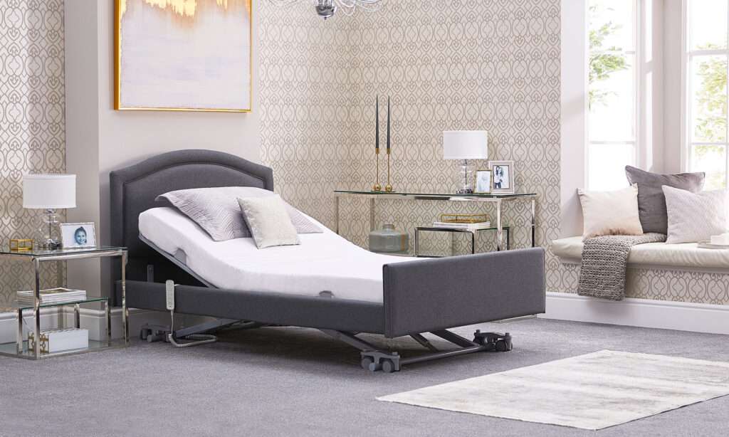 Product shot of the electrically controlled solo comfort profiling mobility recliner bed. The mobility bed is inside a bedroom. This bed is in a reclined position with the wheels visible. There is no person in the bed.