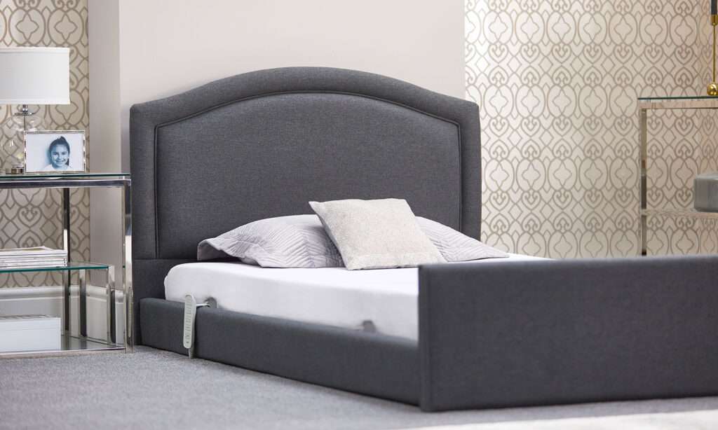 Product shot of the electrically controlled solo comfort profiling mobility recliner bed in a bedroom. The bed is upholstered with a large headboard.