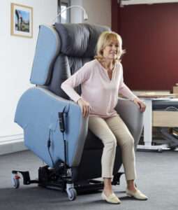 Lifestyle shot of an elderly woman using the ProSpec rise and recline hospital chair as a standing aid. The picture is indoors, and the room is carpeted.