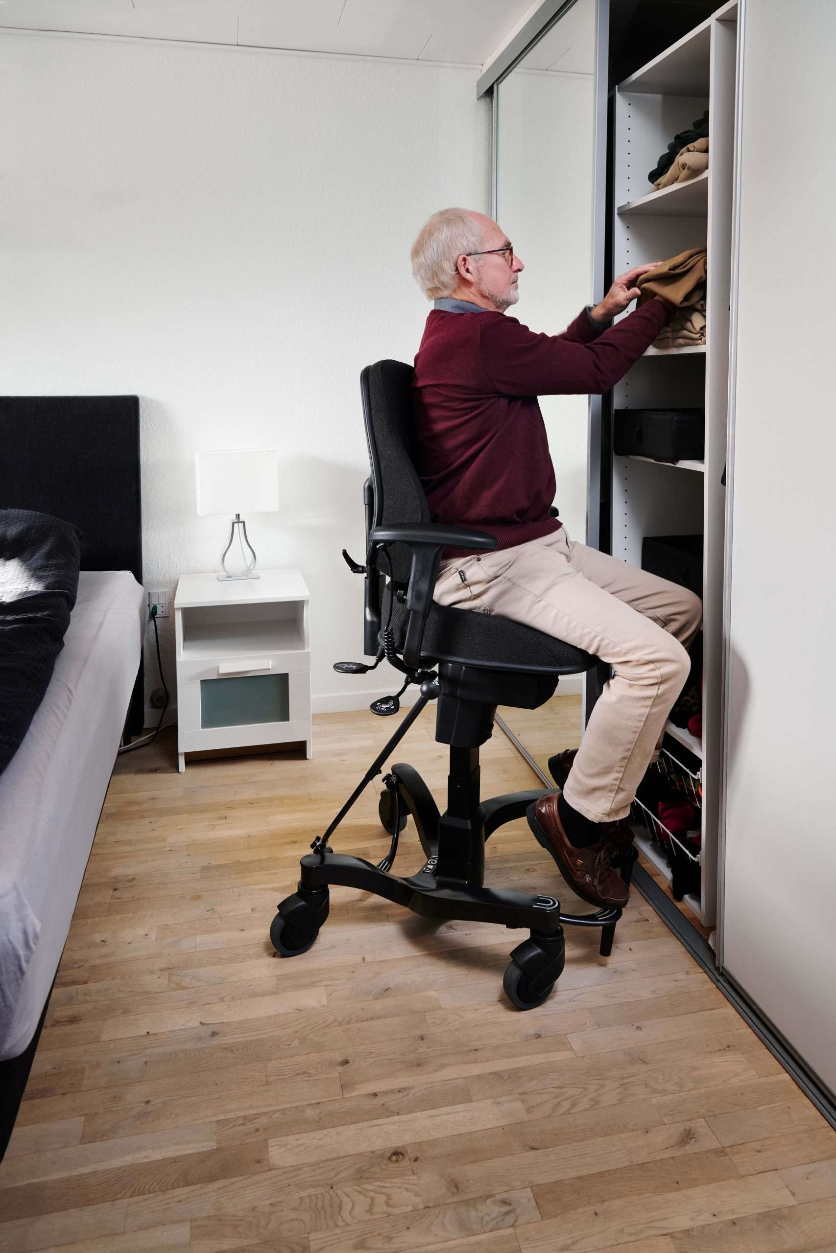 Lifestyle shot of an elderly man in his bedroom sitting on an elevated Vela 700 Tango Activity chair using the raised height to store clothes.