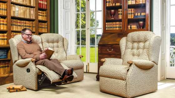 Elderly man in a living room sitting in an orthopaedic recliner chair reading a book. There is an empty orthopaedic recliner chair chair opposite him. A bookshelf is behind both chairs.
