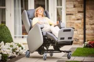 Elderly woman sitting in the dual motor, tilt in space Lento care chair with wheels. She is outdoors.