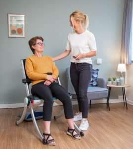 Elderly woman in a sitting position after using the Raizer lift chair. The falls assistant is a younger woman. They are both indoors.