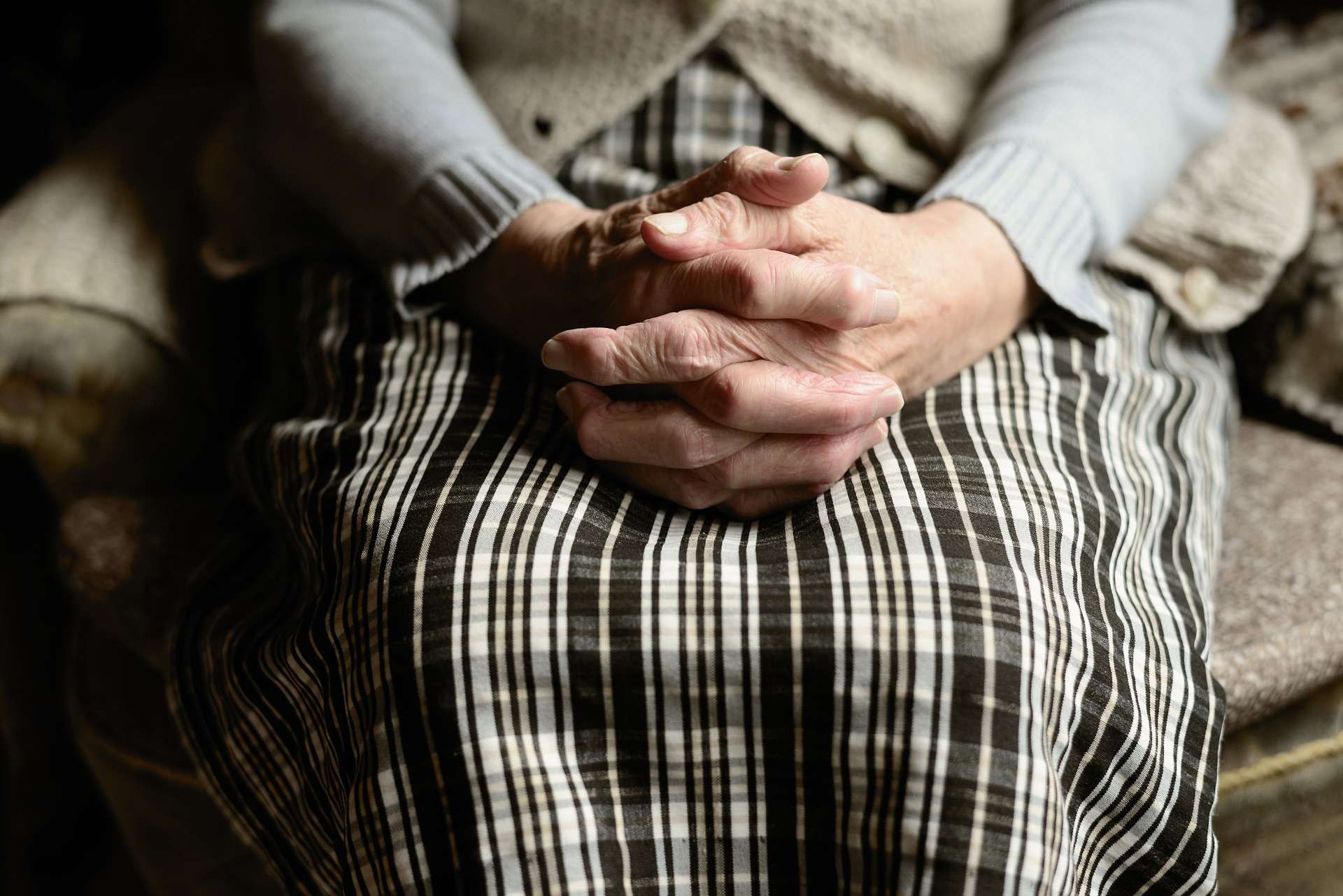 An elderly person sat with their hands clasped on the edge of a bed.