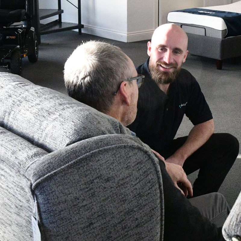 Man sat in a riser recliner chair, speaking to a salesman in the Vivid Care Harrogate mobility shop showroom.
