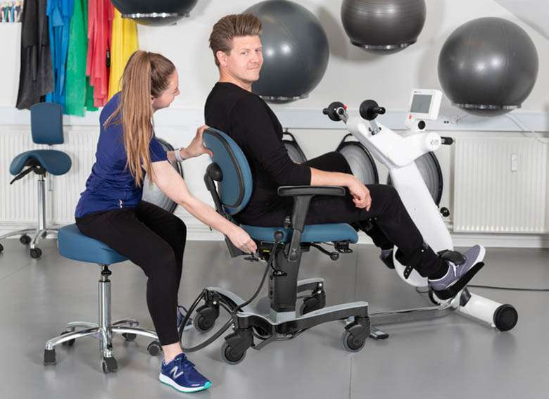 Man in a rehabilitation centre using a Vela exercise chair and a hand bike. A nurse is helping to adjust the chair.