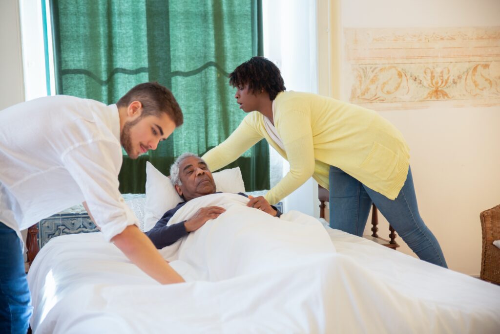Elderly man being cared for bed by two nursing attendants. One is male, the other is female.