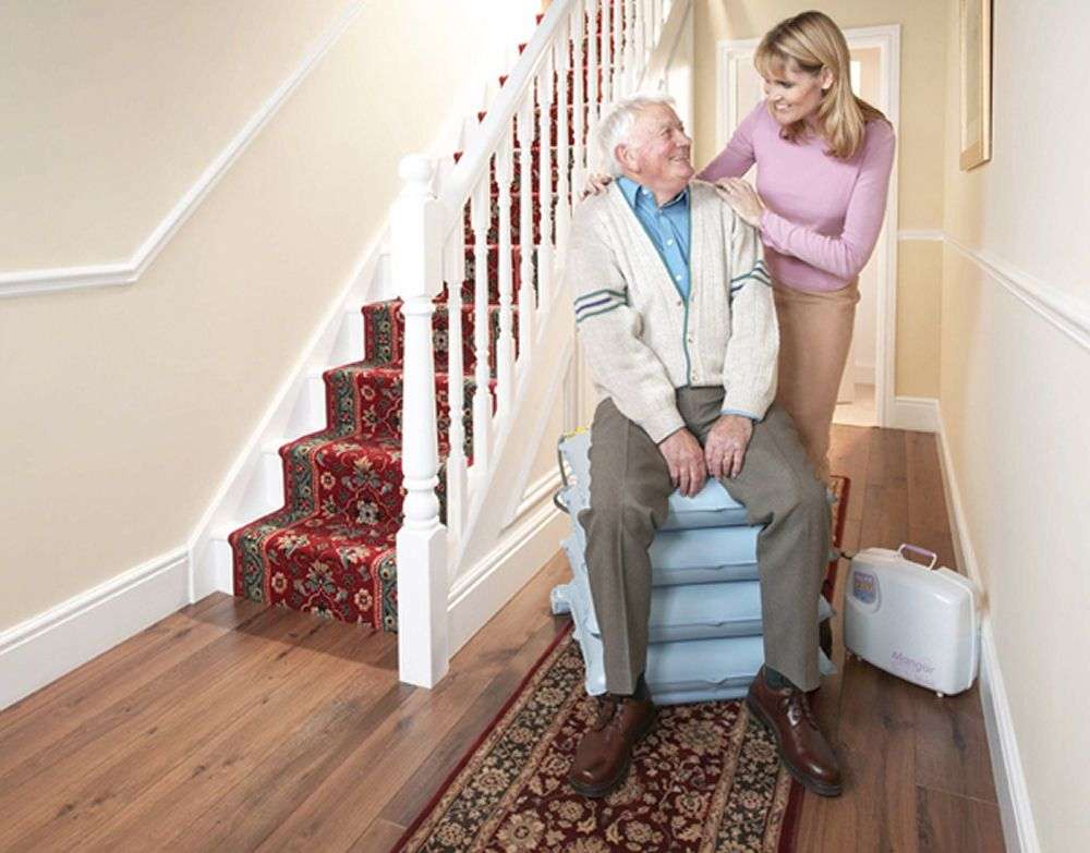 Elderly man using a mangar lifting cushion with the help of a younger woman. The are both positioned next to a staitcase.