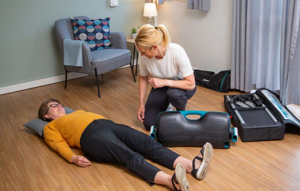 Younger nurse using a raizer 2 electric lifting chair to pick up a fallen older lady from the floor. They are both indoors.