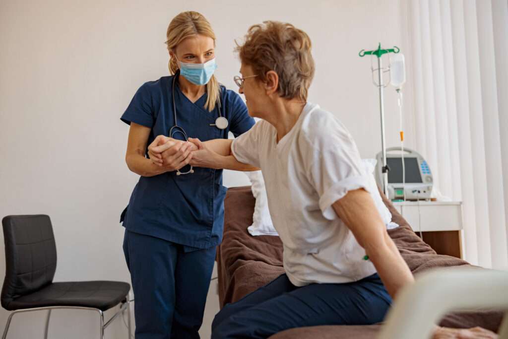 Female nurse in mask helping patient to get up from bed in the ward at hospital. High quality photo