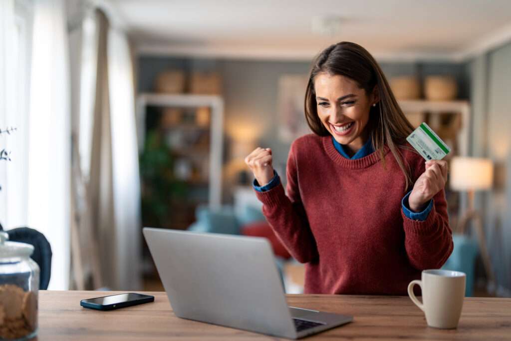 Enthusiastic charming millennial woman feeling excited looking at laptop screen holding credit card, raising fist in yes gesture satisfied with online payment possibilities, rejoicing approved loan.
